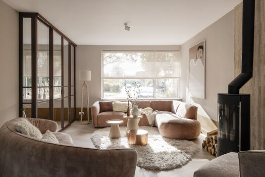 Warme woonkamer met roze bank, bruine fauteuil, open haard, groot kunstwerk en glazen scheidingswand