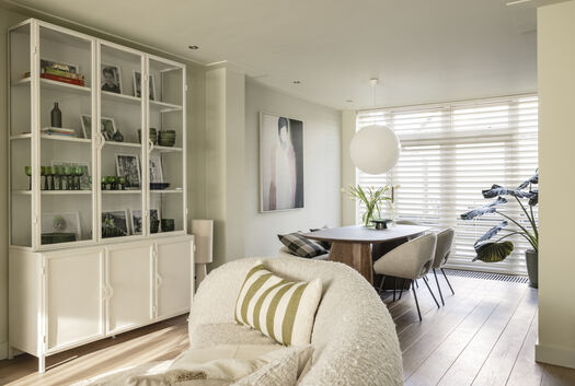 Woonruimte met witte boekenkast, beige fauteuil met gestreept kussen, eethoek en abstract kunstwerk