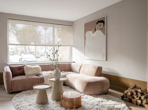 Moderne woonkamer met roze fluwelen hoekbank, ronde tafels, wit kleed en abstract portret aan muur.