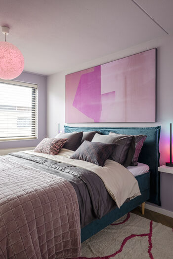 Slaapkamer met groot roze abstract kunstwerk, fluwelen blauwgroen bed en kleurrijke verlichting