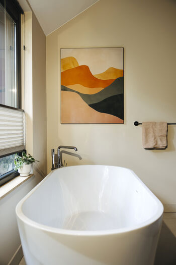 Moderne badkamer met vrijstaand wit bad, abstract berglandschap kunstwerk en natuurlijk licht.