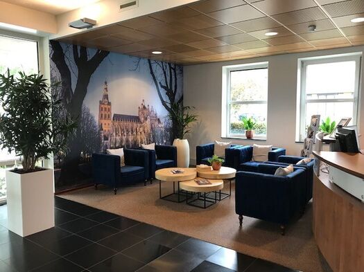 Wachtkamer met blauwe fauteuils, ronde tafels en muurdecoratie met stadsgezicht.