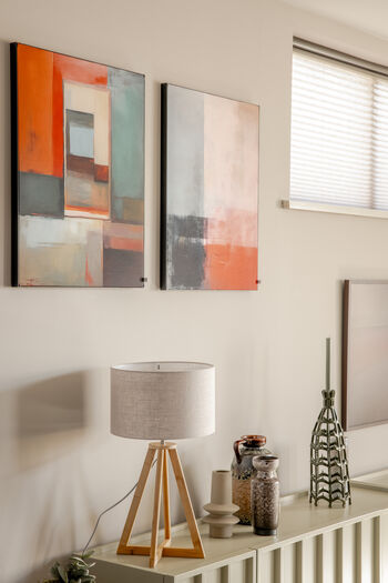 Minimalistisch interieur met twee abstracte schilderijen in oranje en grijstinten boven houten lamp