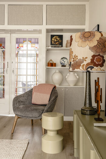 Modern interieur met grijze fauteuil, crème poef, ingebouwde kast en vintage bloemen wanddecoratie
